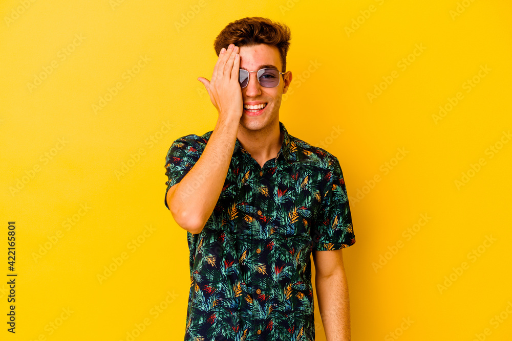 Young caucasian man wearing a Hawaiian shirt isolated on yellow background having fun covering half of face with palm. Young caucasian man wearing a Hawaiian shirt isolated on yellow background having fun covering half of face with palm.