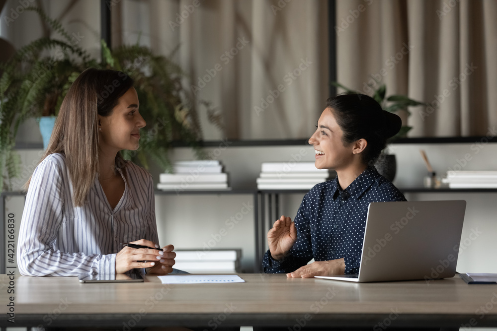 Smiling young multiethnic female colleagues coworkers sit at table in office talk brainstorm over company business project. Happy diverse women employees speak discuss ideas using laptop. Smiling young multiethnic female colleagues coworkers sit at table in office talk brainstorm over company business project. Happy diverse women employees speak discuss ideas using laptop.