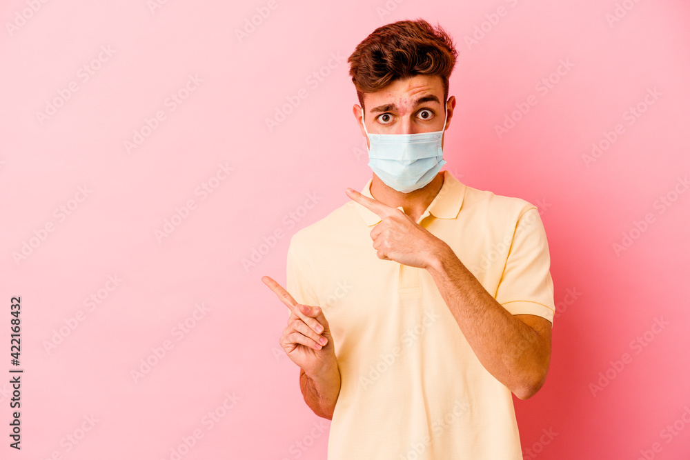 Young caucasian man wearing a protection for coronavirus isolated on pink background shocked pointing with index fingers to a copy space.