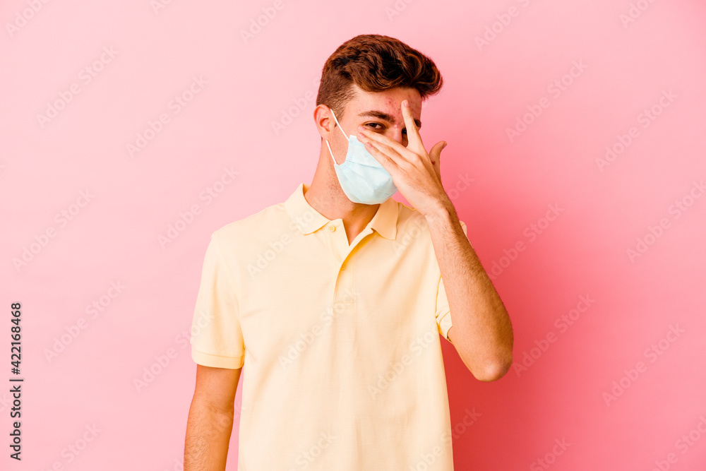 Young caucasian man wearing a protection for coronavirus isolated on pink background blink at the camera through fingers, embarrassed covering face. Young caucasian man wearing a protection for coronavirus isolated on pink background blink at the camera through fingers, embarrassed covering face.