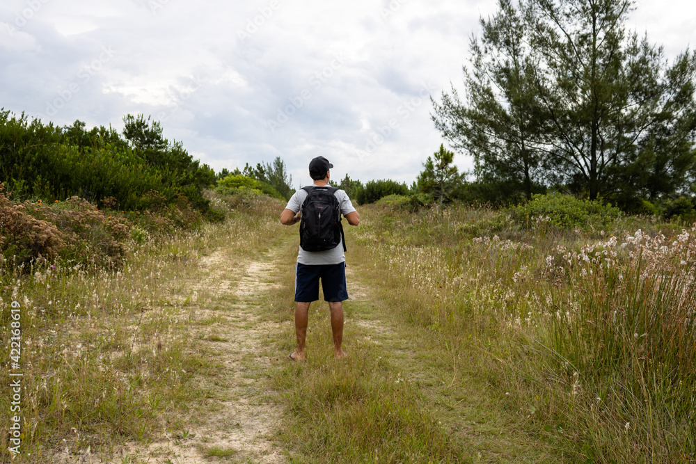 Brunette man with a backpack and cap wearing a gray t-shirt and flip flops on a path next to a secluded beach looking up at the sky on a cloudy, rainy day. Reference to vacation trip. Brunette man with a backpack and cap wearing a gray t-shirt and flip flops on a path next to a secluded beach looking up at the sky on a cloudy, rainy day. Reference to vacation trip.