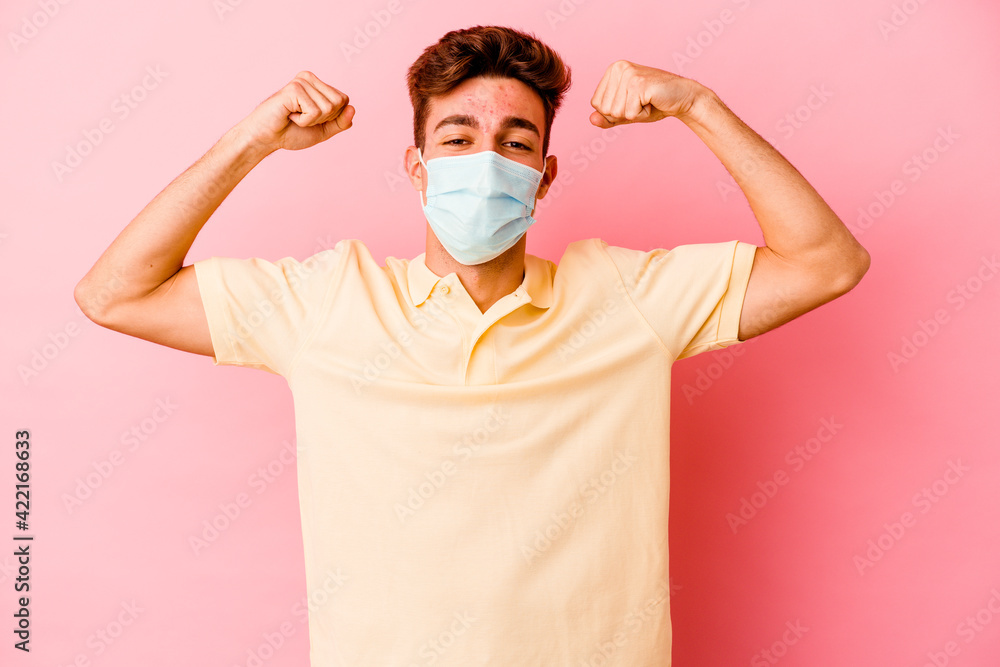 Young caucasian man wearing a protection for coronavirus isolated on pink background showing strength gesture with arms, symbol of feminine power