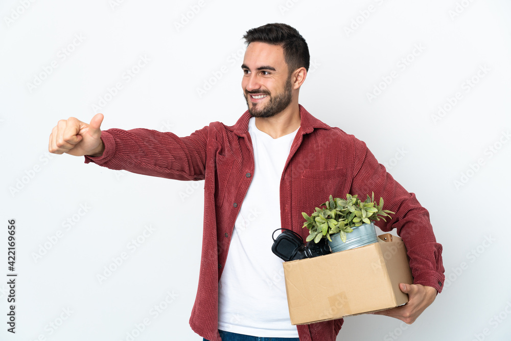Young caucasian man making a move while picking up a box full of things isolated on white background giving a thumbs up gesture Young caucasian man making a move while picking up a box full of things isolated on white background giving a thumbs up gesture
