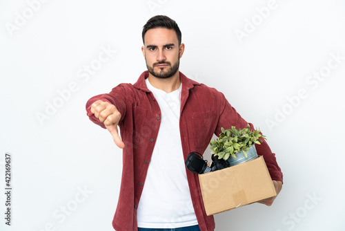 Young caucasian man making a move while picking up a box full of things isolated on white background showing thumb down with negative expression