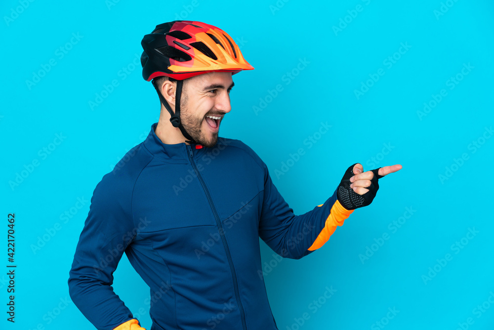 Young cyclist man isolated on blue background pointing finger to the side and presenting a product - obrazy, fototapety, plakaty 