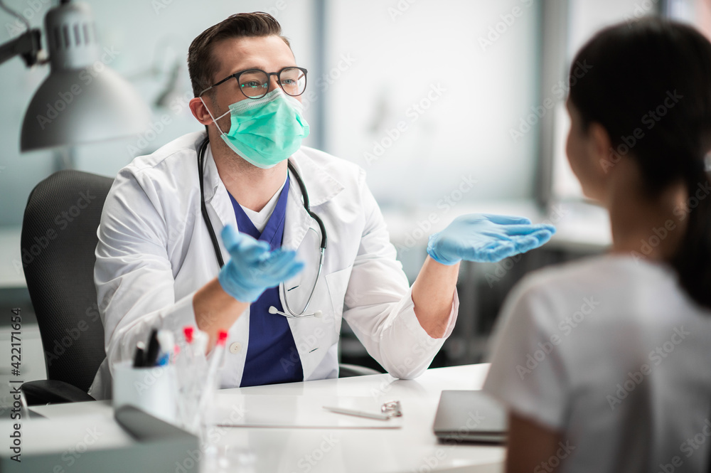 A young doctor sees a female patient at a desk in his office, wearing a mask and gloves.