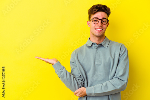 Young caucasian man isolated on yellow background showing a copy space on a palm and holding another hand on waist.