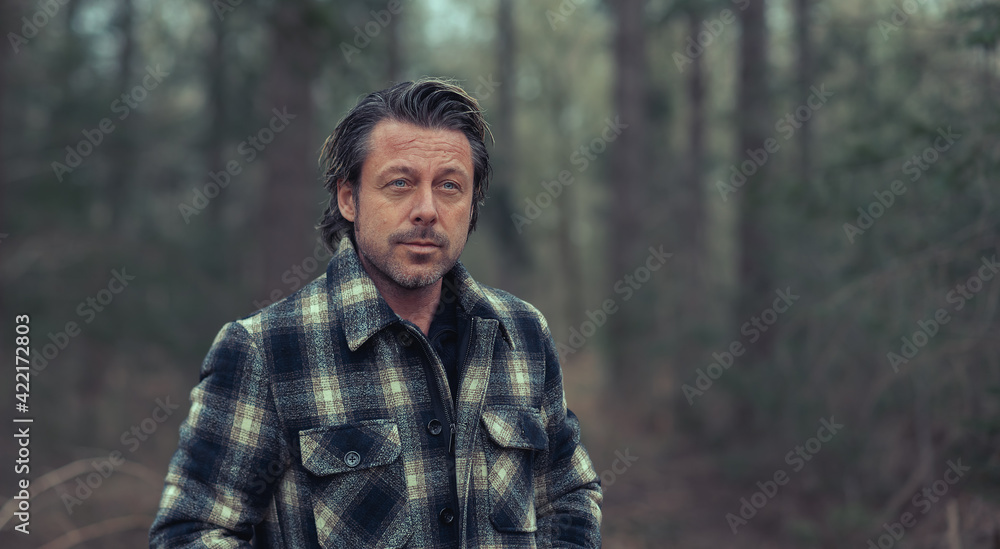 Blonde man in checkered coat standing in the woods.