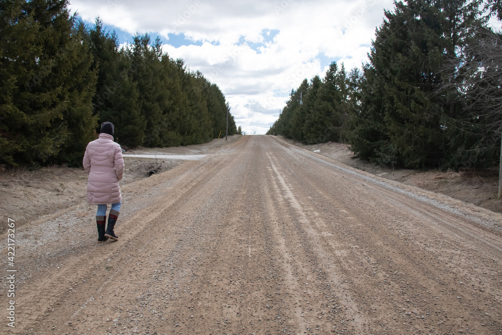 A woman wearing a pink puff parka and a black toque and jeans walks down an old dirt road in the countryside lined with coniferous spruce trees in March, 2021, Ontario Canada.
