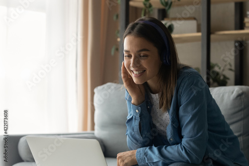 Close up smiling woman wearing headphones chatting  using laptop  looking at computer screen  sitting on couch at home  happy young female making video call to relatives or friends  talking