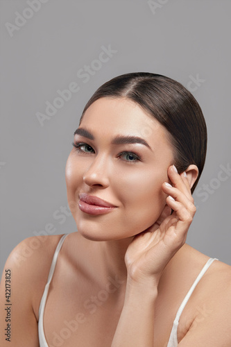 Beauty face. Smiling woman touching healthy skin portrait. Beautiful happy girl model with fresh glowing facial skin and natural makeup on gray background at studio. Skin care concept