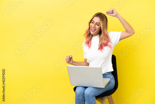 Young woman sitting on a chair with laptop over isolated yellow background celebrating a victory