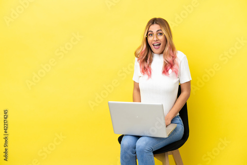 Young woman sitting on a chair with laptop over isolated yellow background with surprise facial expression