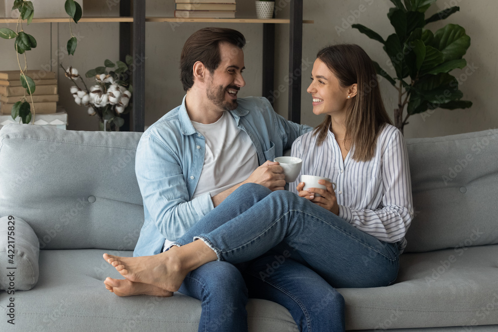 Happy young couple relaxing on cozy couch at home, holding cups, drinking hot beverages, tea or coffee together, laughing man and woman enjoying leisure time, chatting, sharing news, having fun Happy young couple relaxing on cozy couch at home, holding cups, drinking hot beverages, tea or coffee together, laughing man and woman enjoying leisure time, chatting, sharing news, having fun