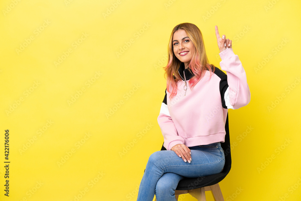 Young woman sitting on a chair over isolated yellow background showing and lifting a finger in sign of the best Young woman sitting on a chair over isolated yellow background showing and lifting a finger in sign of the best