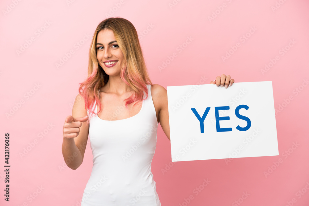 Young woman over isolated pink background holding a placard with text YES and pointing it Young woman over isolated pink background holding a placard with text YES and pointing it