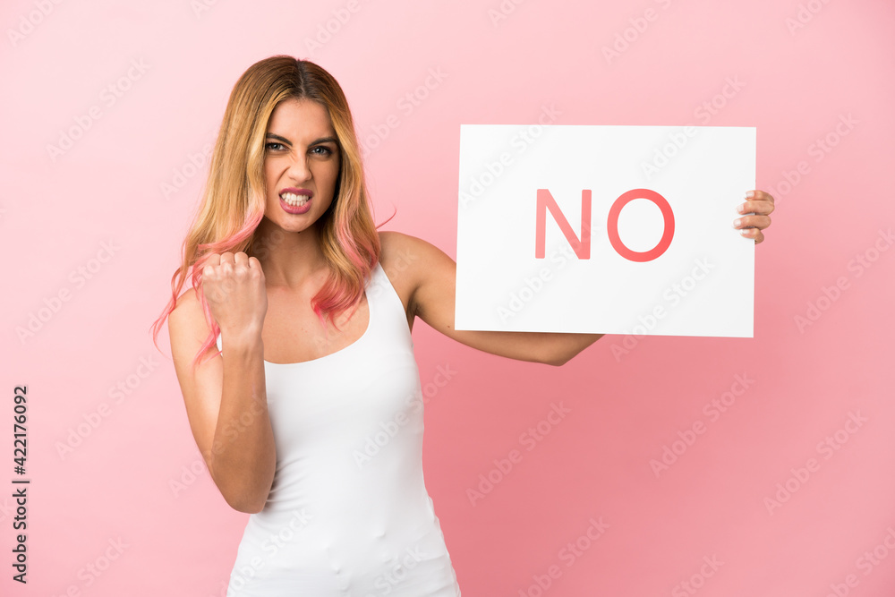 Young woman over isolated pink background holding a placard with text NO and angry Young woman over isolated pink background holding a placard with text NO and angry