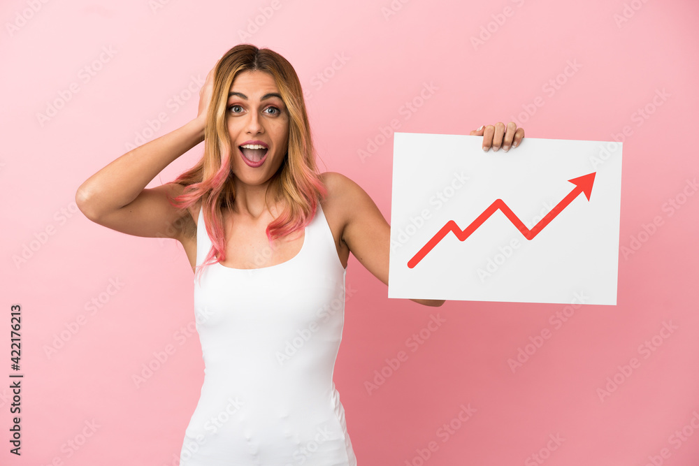 Young woman over isolated pink background holding a sign with a growing statistics arrow symbol with surprised expression Young woman over isolated pink background holding a sign with a growing statistics arrow symbol with surprised expression