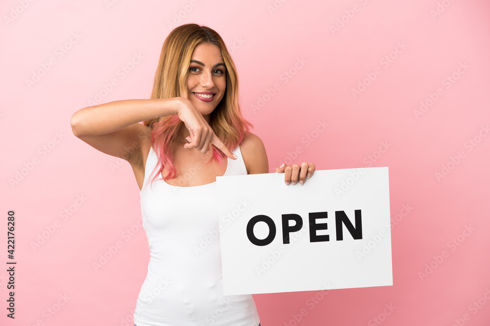 Young woman over isolated pink background holding a placard with text OPEN and pointing it Young woman over isolated pink background holding a placard with text OPEN and pointing it