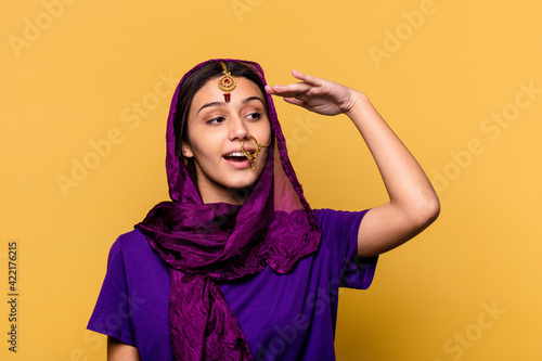 Young Indian woman wearing a traditional sari clothes isolated on yellow background looking far away keeping hand on forehead. © Asier
