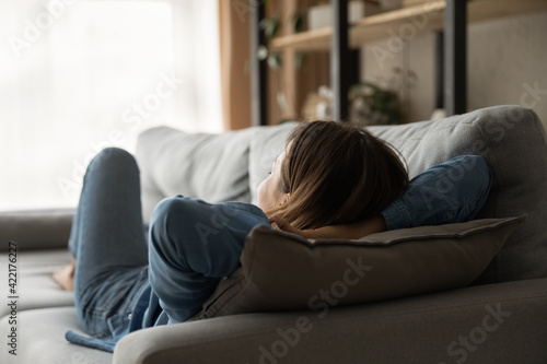 Rear view calm woman relaxing, lying with hands behind head on comfortable couch at home, peaceful young female enjoying lazy weekend, resting, taking nap, sleeping or daydreaming in living room