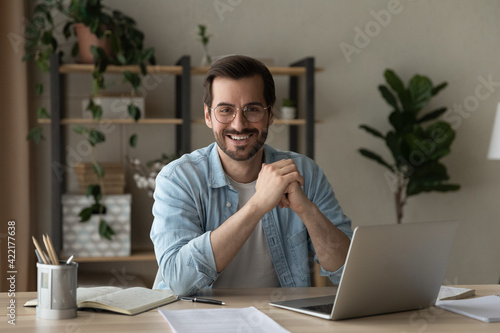 Head shot portrait smiling confident businessman wearing glasses sitting at table with laptop and documents, looking at camera, successful happy entrepreneur or student posing at workplace