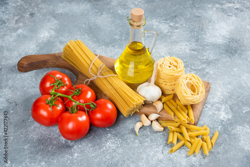 Assortment of raw pasta, olive oil and tomatoes on wooden board