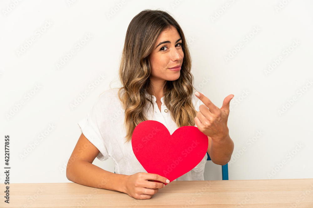Young caucasian woman holding a valentines day heart isolated pointing with finger at you as if inviting come closer. Young caucasian woman holding a valentines day heart isolated pointing with finger at you as if inviting come closer.