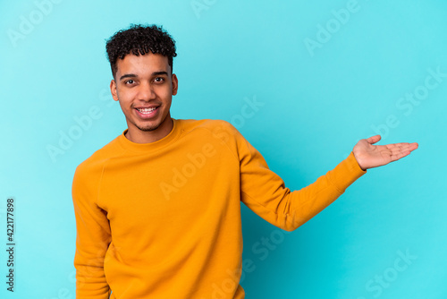 Young african american curly man isolated on blue showing a copy space on a palm and holding another hand on waist.