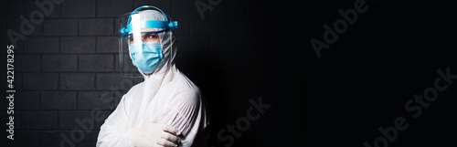 Studio panoramic banner; portrait of a doctor wearing PPE suit against coronavirus and covid-19, on the background of black brick wall with copy space. Pandemic concept.