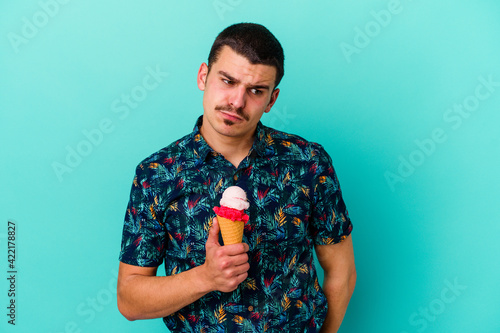 Young caucasian man eating an ice cream isolated on blue background confused, feels doubtful and unsure.