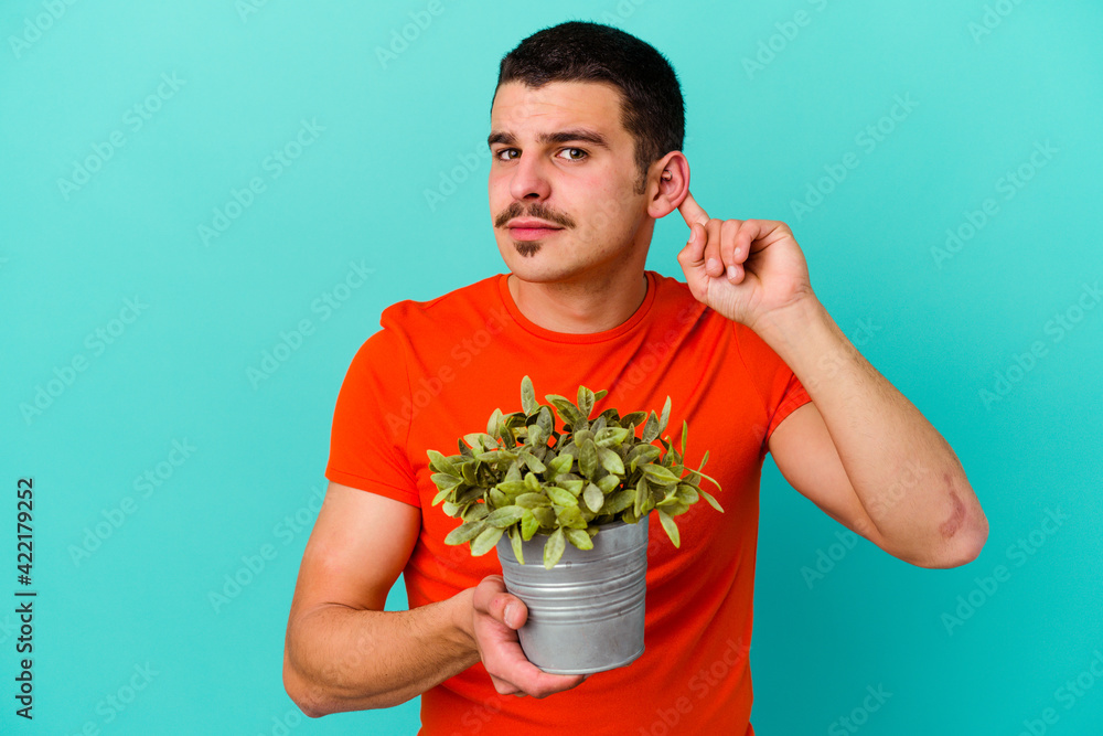 Young caucasian man holding a leaf isolated on blue background trying to listening a gossip. Young caucasian man holding a leaf isolated on blue background trying to listening a gossip.