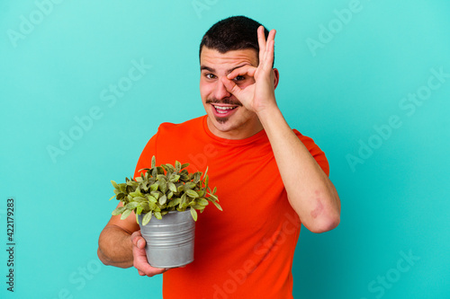 Young caucasian man holding a leaf isolated on blue background excited keeping ok gesture on eye.