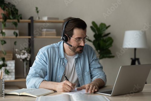 Focused young man wearing headphones and glasses using laptop, watching webinar, writing taking notes, looking at screen, student listening to lecture, studying online at home, sitting at desk