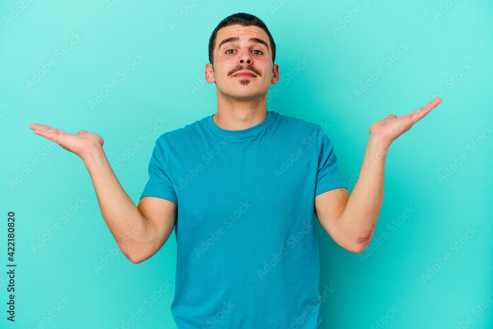 Young caucasian man isolated on blue background confused and doubtful shrugging shoulders to hold a copy space. Young caucasian man isolated on blue background confused and doubtful shrugging shoulders to hold a copy space.
