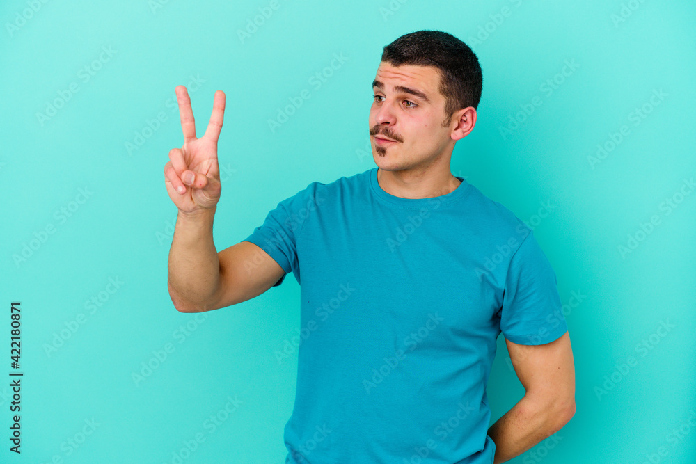 Young caucasian man isolated on blue background joyful and carefree showing a peace symbol with fingers. Young caucasian man isolated on blue background joyful and carefree showing a peace symbol with fingers.