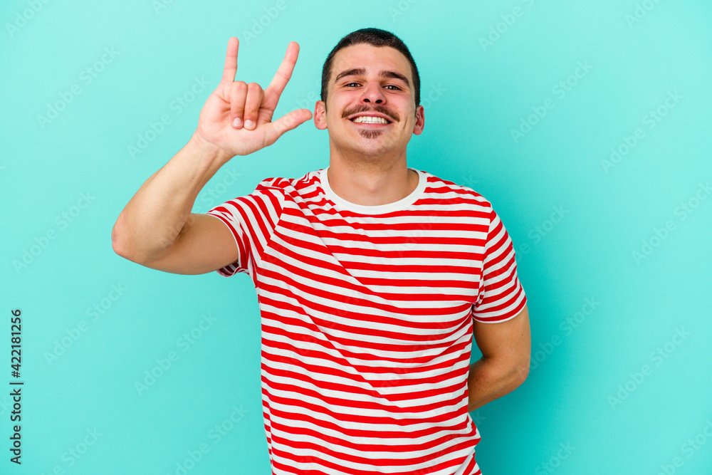 Young caucasian man isolated on blue background showing rock gesture with fingers Young caucasian man isolated on blue background showing rock gesture with fingers