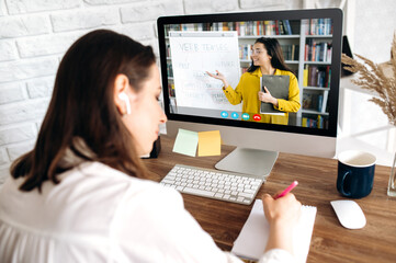 Online education by video conference. Smart girl, studying remotely, taking notes, on the computer screen the teacher conducts an online lesson showing information on a flipchart, studying from home