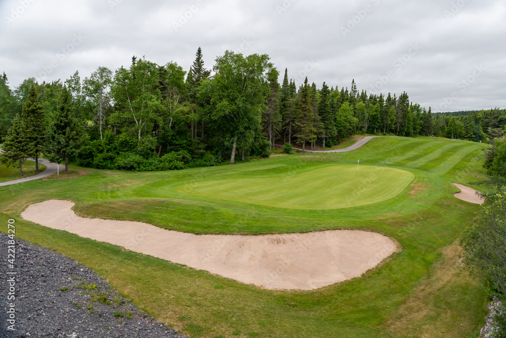 A hole on a golf course with green grass, a sand trap in the shape of a kidney, and lush green trees near the fairground. The sky is cloudy with heavy white and grey clouds. 