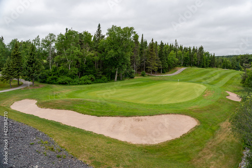 A hole on a golf course with green grass, a sand trap in the shape of a kidney, and lush green trees near the fairground. The sky is cloudy with heavy white and grey clouds. 
