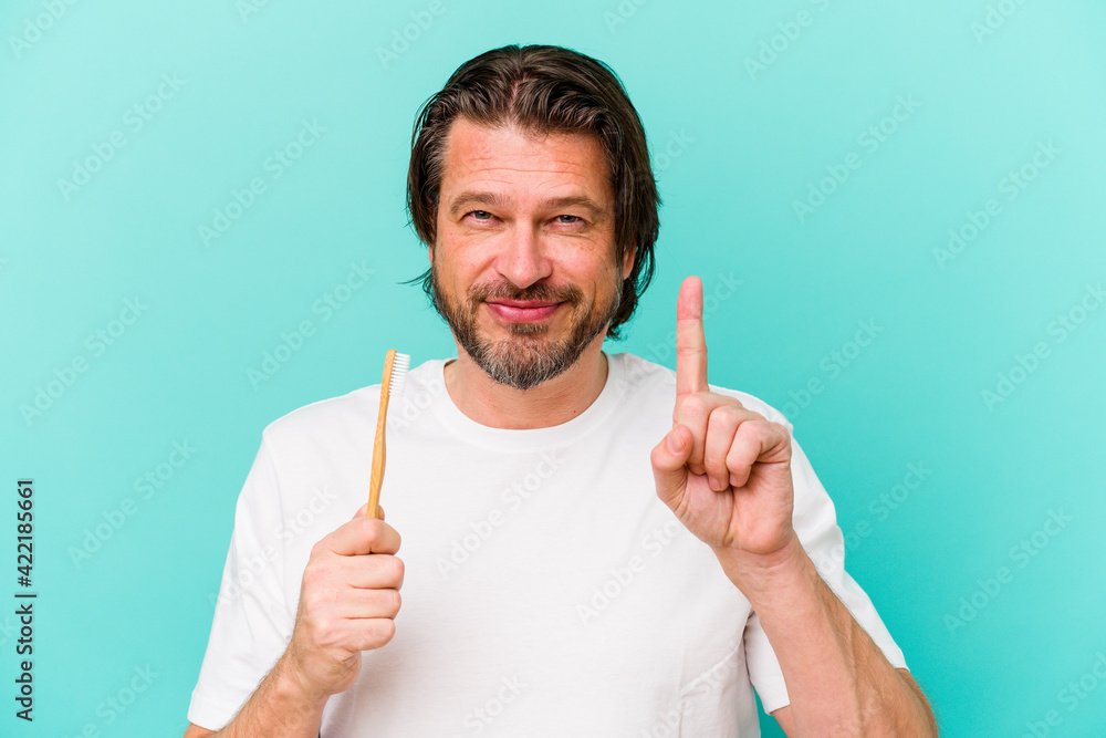 Middle age dutch man holding a toothbrush isolated on blue background showing number one with finger. Middle age dutch man holding a toothbrush isolated on blue background showing number one with finger.