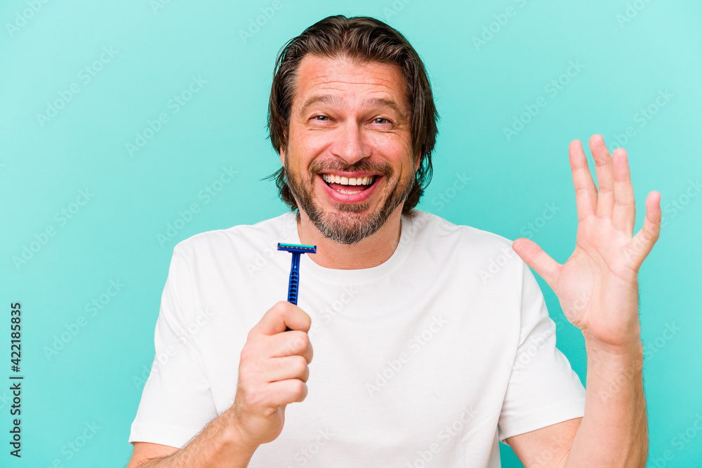 Middle age dutch man holding a razor blade isolated on blue background receiving a pleasant surprise, excited and raising hands. Middle age dutch man holding a razor blade isolated on blue background receiving a pleasant surprise, excited and raising hands.