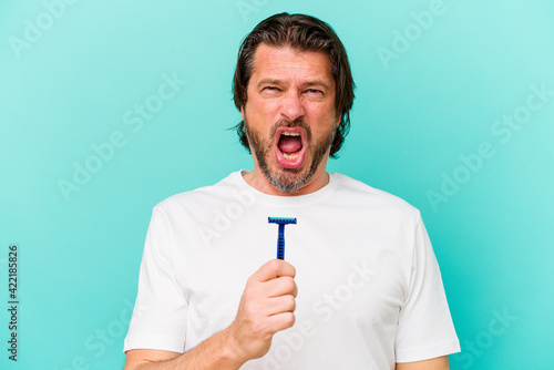 Middle age dutch man holding a razor blade isolated on blue background screaming very angry and aggressive.