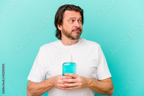 Middle age dutch man sitting holding a mouthwash isolated on blue background confused, feels doubtful and unsure.