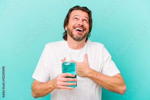 Middle age dutch man sitting holding a mouthwash isolated on blue background laughs out loudly keeping hand on chest.