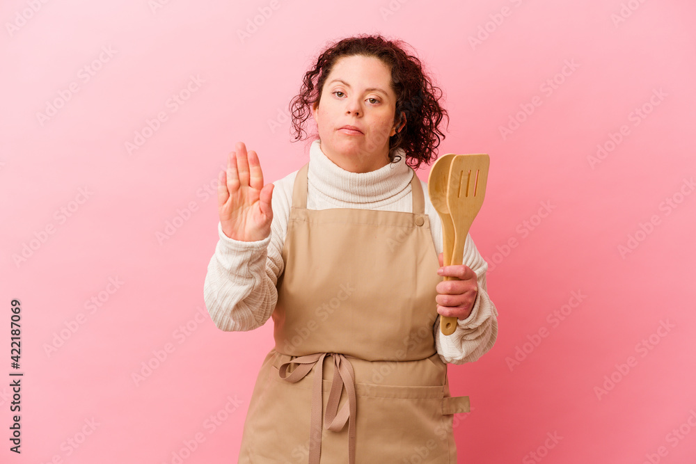 Woman with Down syndrome cooking at home isolated on pink background standing with outstretched hand showing stop sign, preventing you.