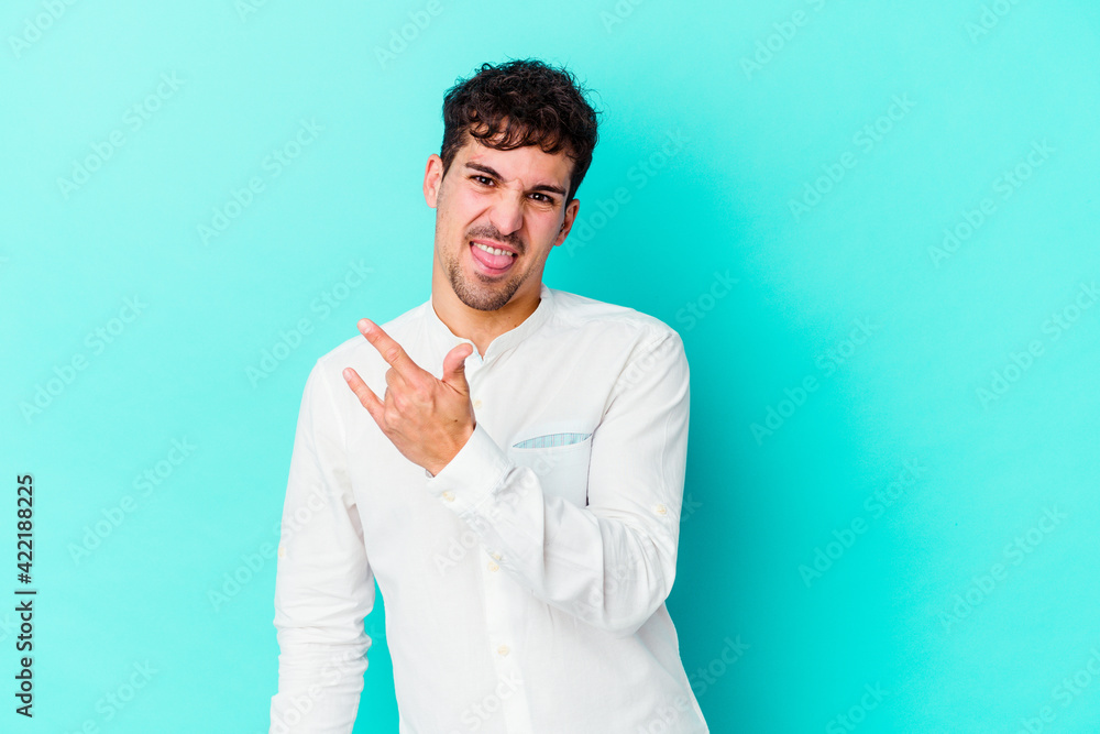 Young caucasian man isolated on blue background showing rock gesture with fingers Young caucasian man isolated on blue background showing rock gesture with fingers