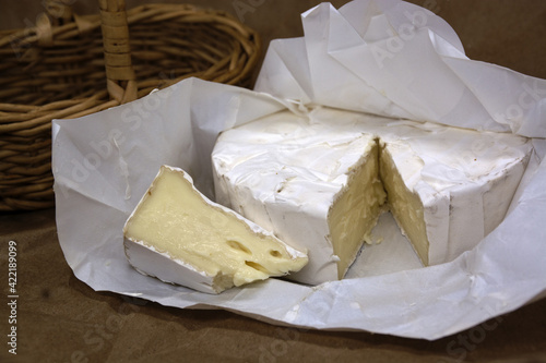 Fresh brie cheese and one slice on white paper. Camembert cheese with wooden basket on background.