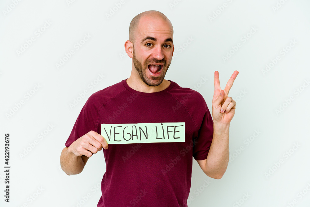 Young caucasian bald man holding a Vegan life placard isolated on purple background joyful and carefree showing a peace symbol with fingers. Young caucasian bald man holding a Vegan life placard isolated on purple background joyful and carefree showing a peace symbol with fingers.