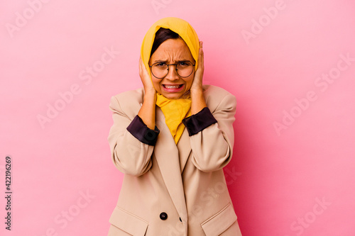 Young business muslim woman isolated on pink background covering ears with hands trying not to hear too loud sound.